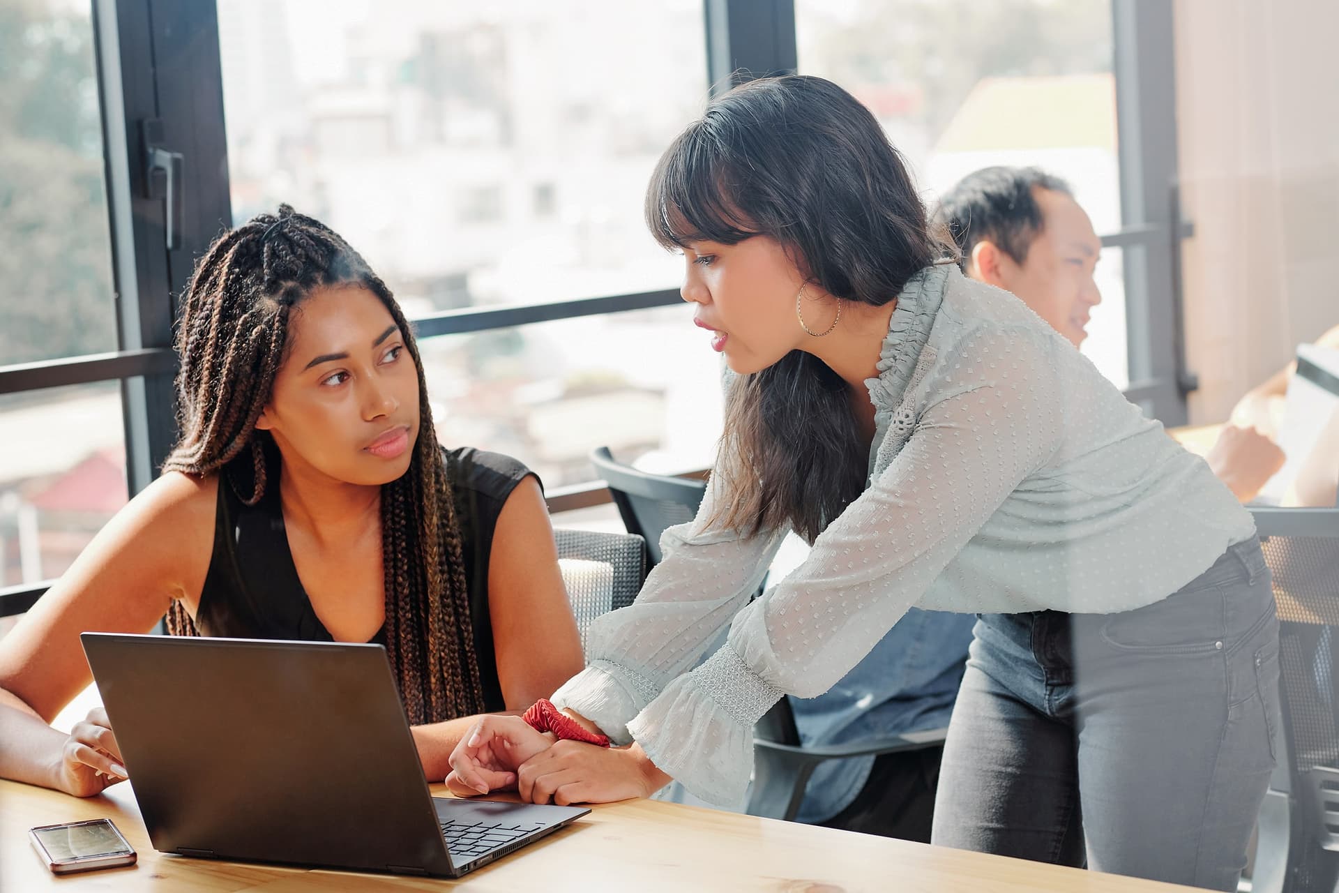 A woman pointing at a laptop while a seated colleague listens, by a window