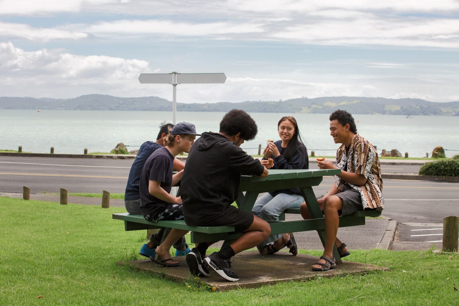 Five young people sharing food at a picnic table across the road from the water