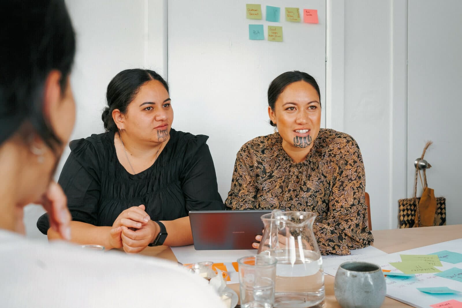 Two women, both with moko kauae, talking at a table, sticky notes on the wall behind