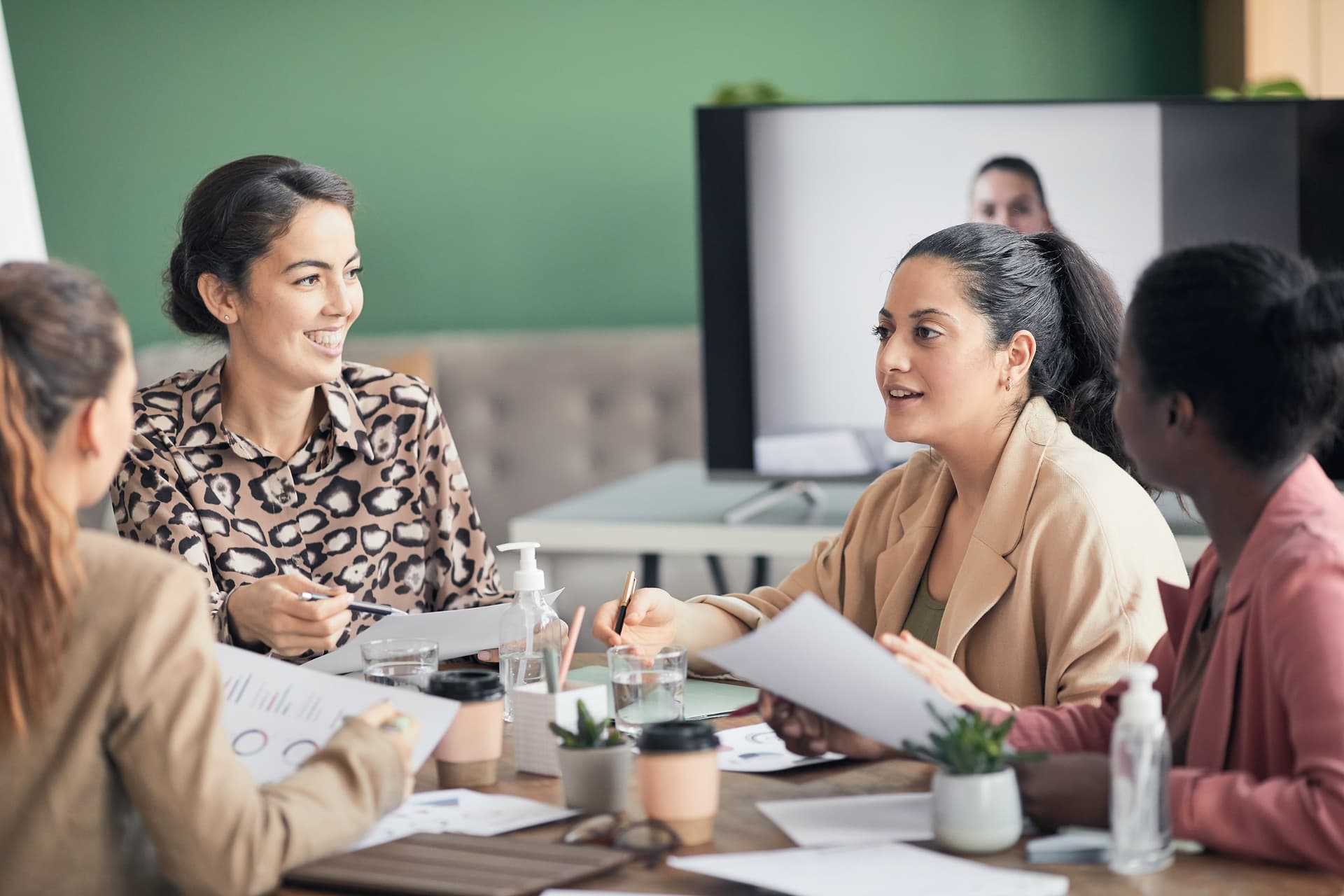 Four women discussing printed charts at a table, a colleague on a video screen behind