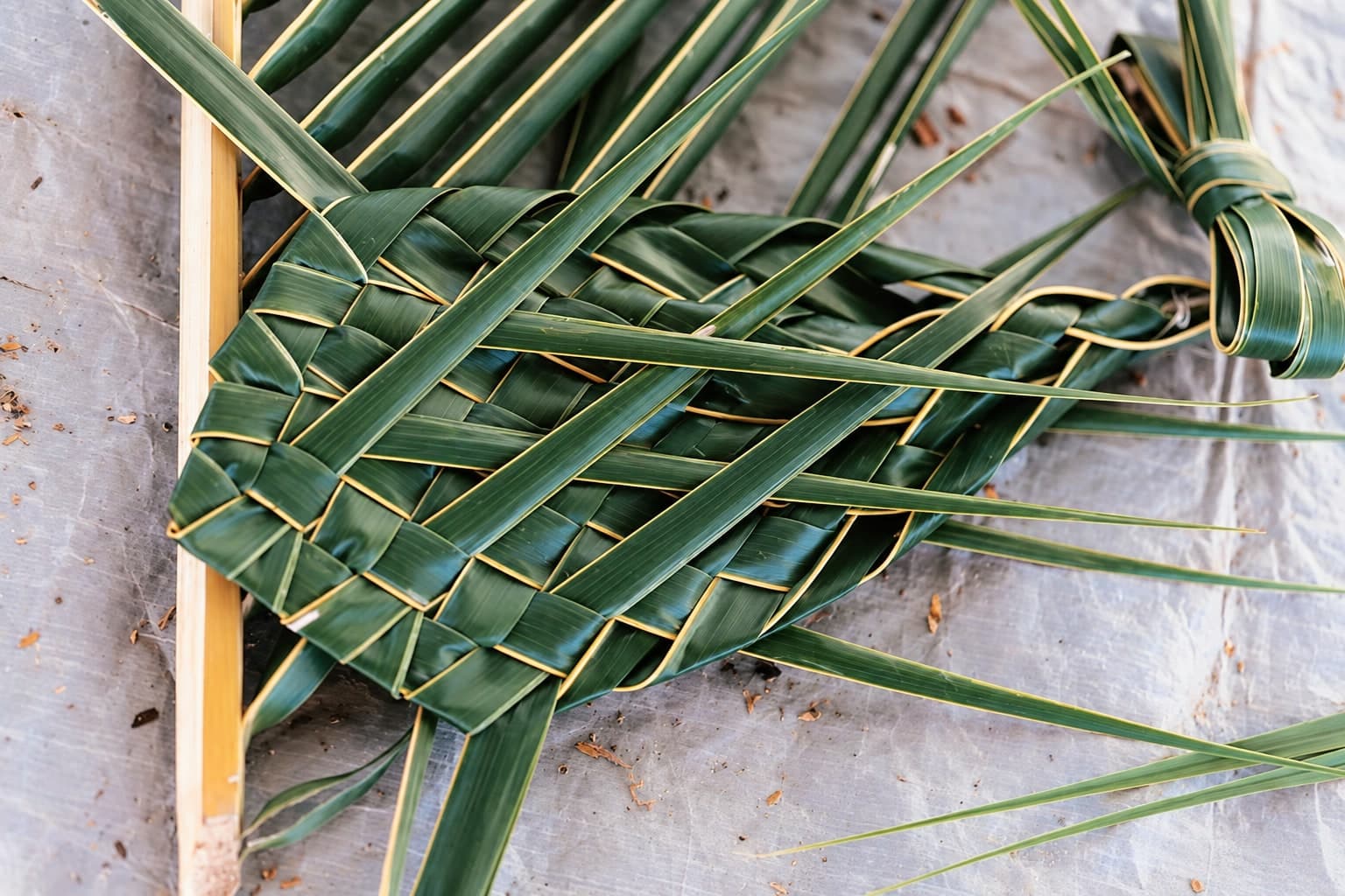 Green harakeke strips partly woven into a mat on a pale grey mottled surface.