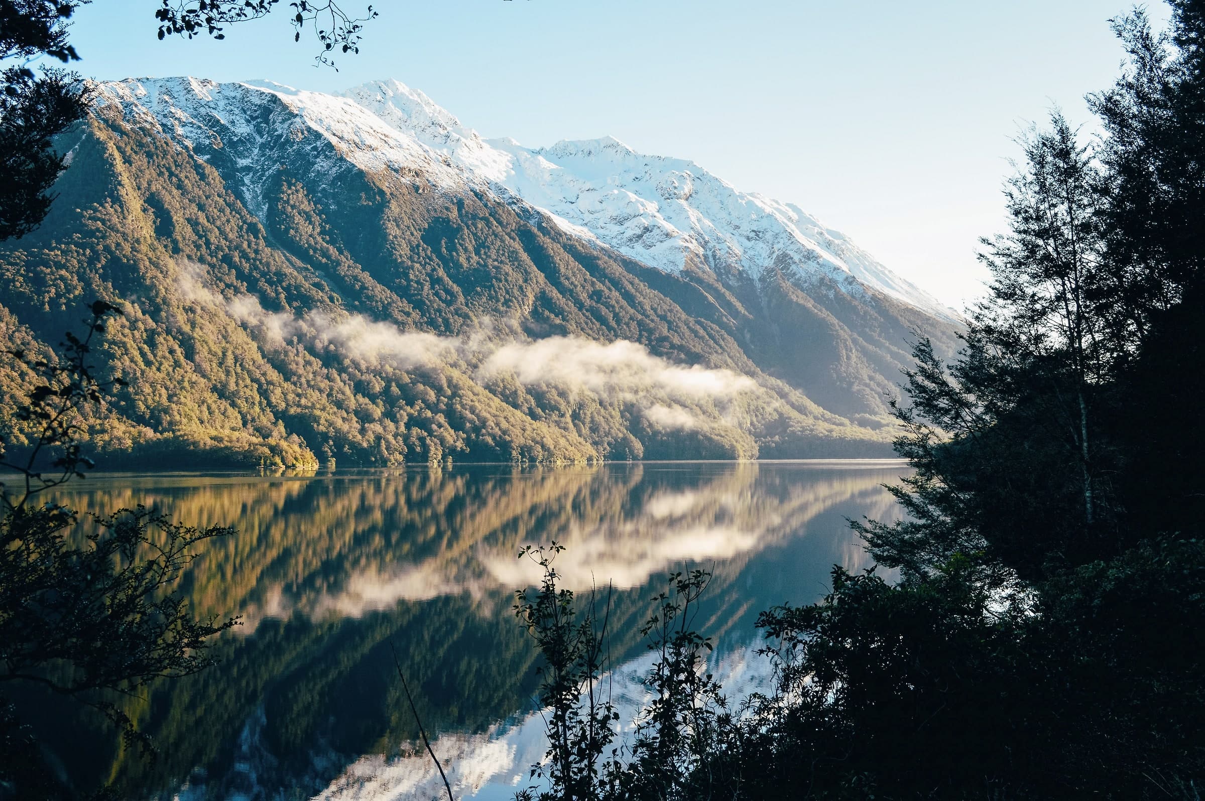 A still lake reflecting bush-covered mountains, mist low on the hills