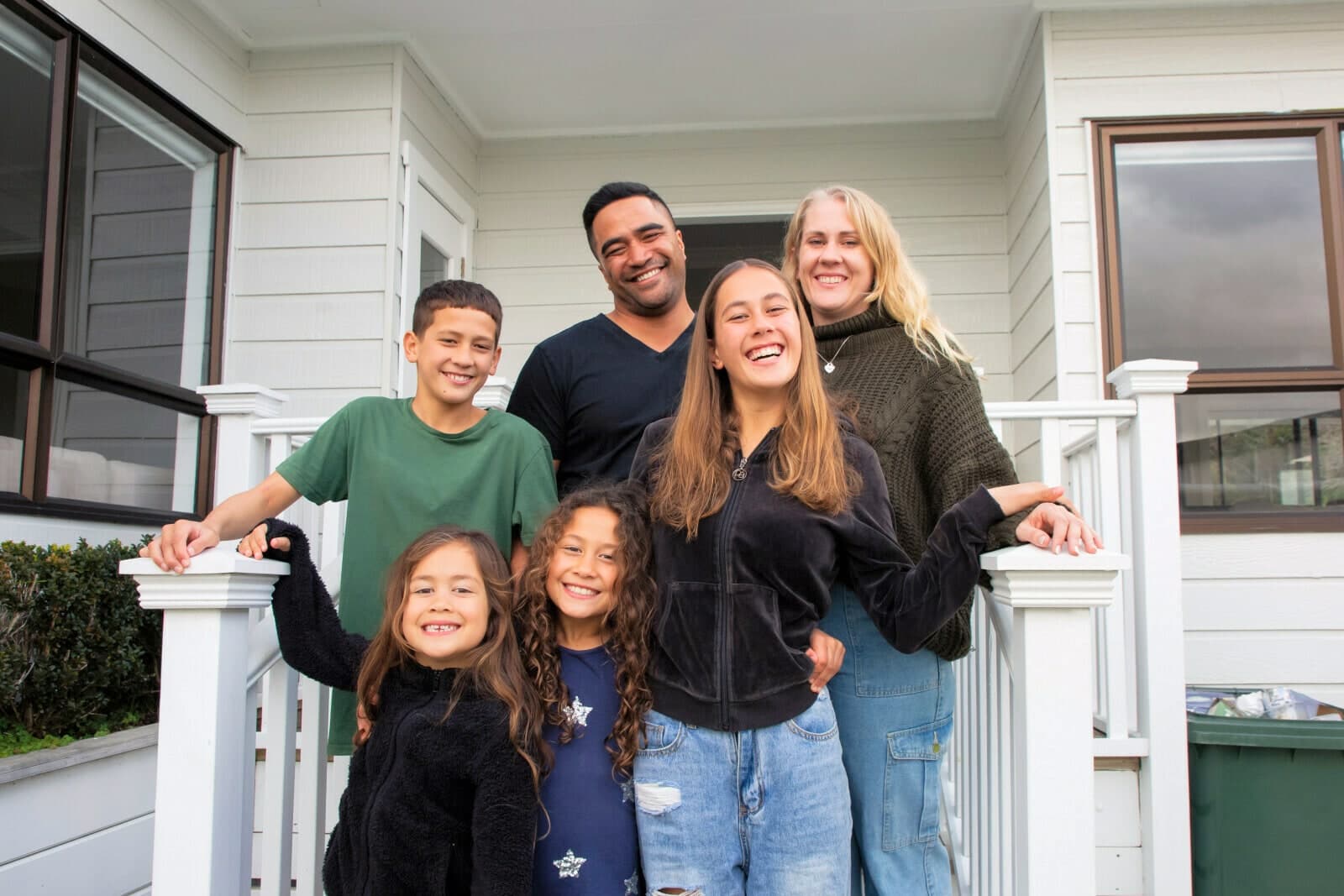 A whānau smiling on the front steps of a weatherboard house